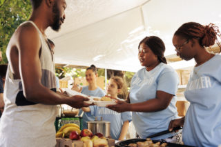 Multiethnic female volunteers serving free food to the homeless man at an outdoor food bank. Non-profit group offers humanitarian aid and fosters community, helping the hungry and less privileged.